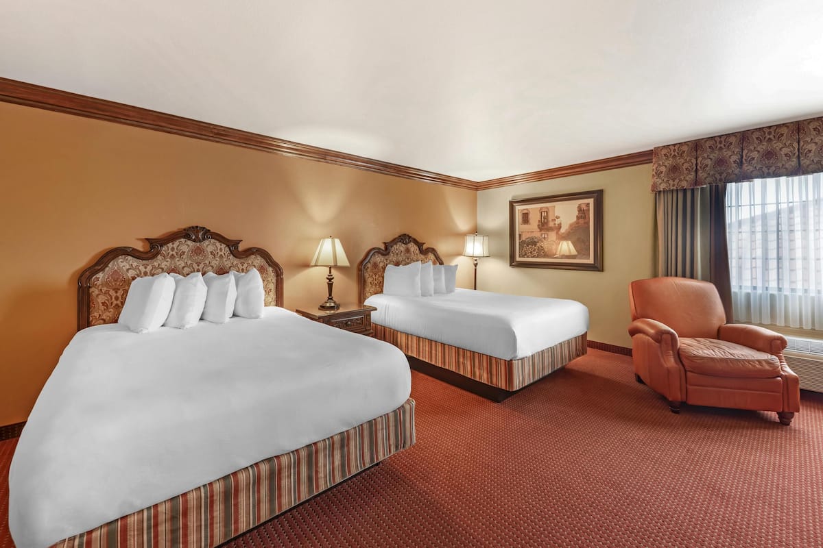 Quarto de hotel com duas camas, uma cadeira, um quadro e uma janela com cortinas.