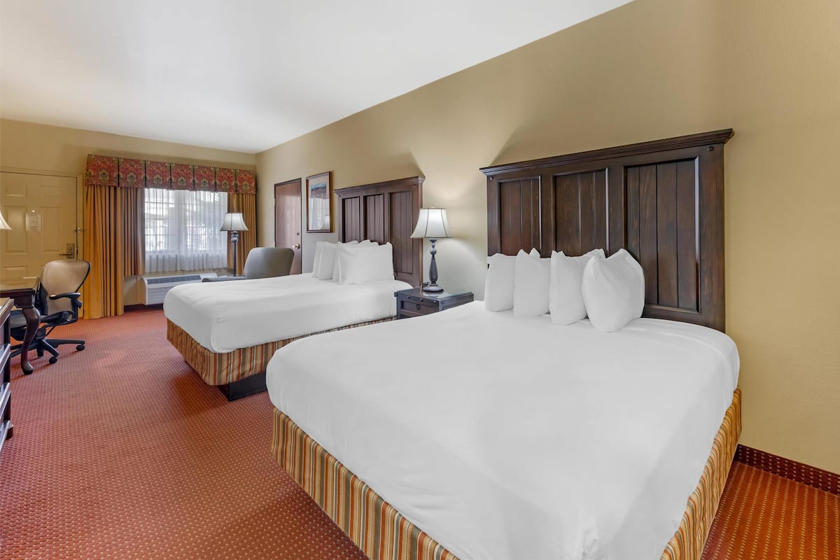 Quarto de hotel com duas camas, uma mesa, uma cadeira e uma janela com cortinas.