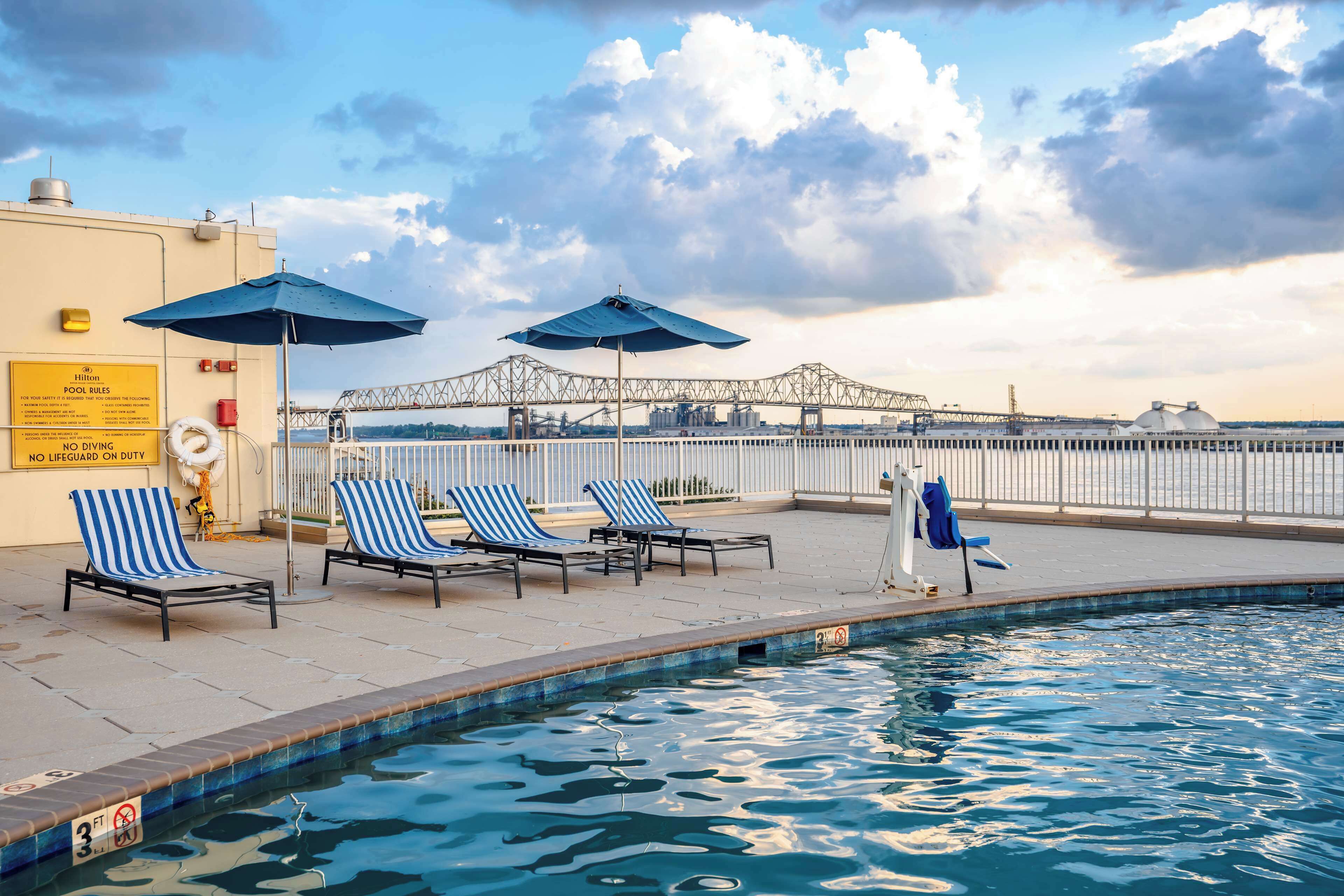 A pool area with lounge chairs and umbrellas, a sign indicating no diving, and a view of a bridge in the background.