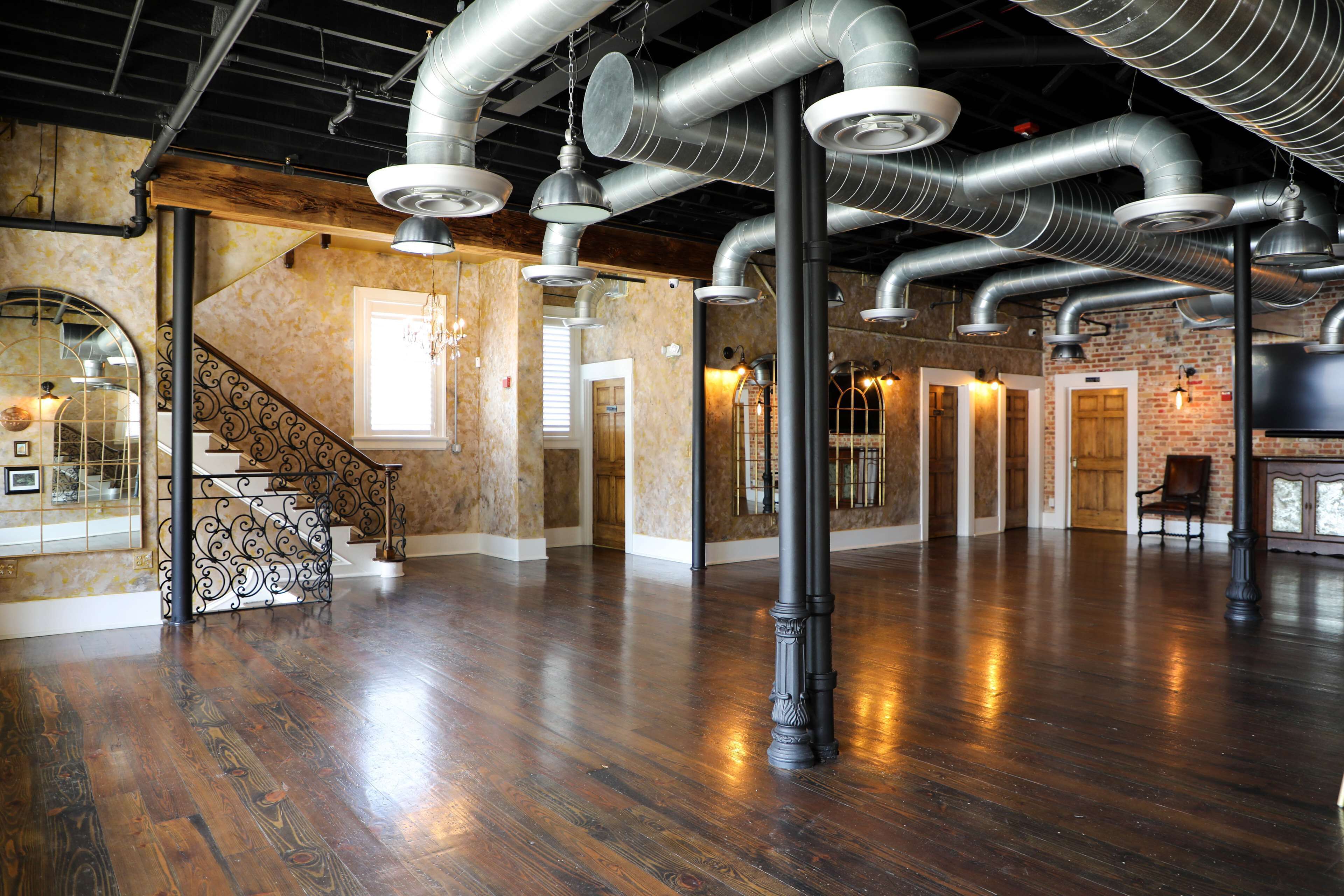 A spacious room with polished wooden floors, exposed ductwork, and a staircase with ornate railings.