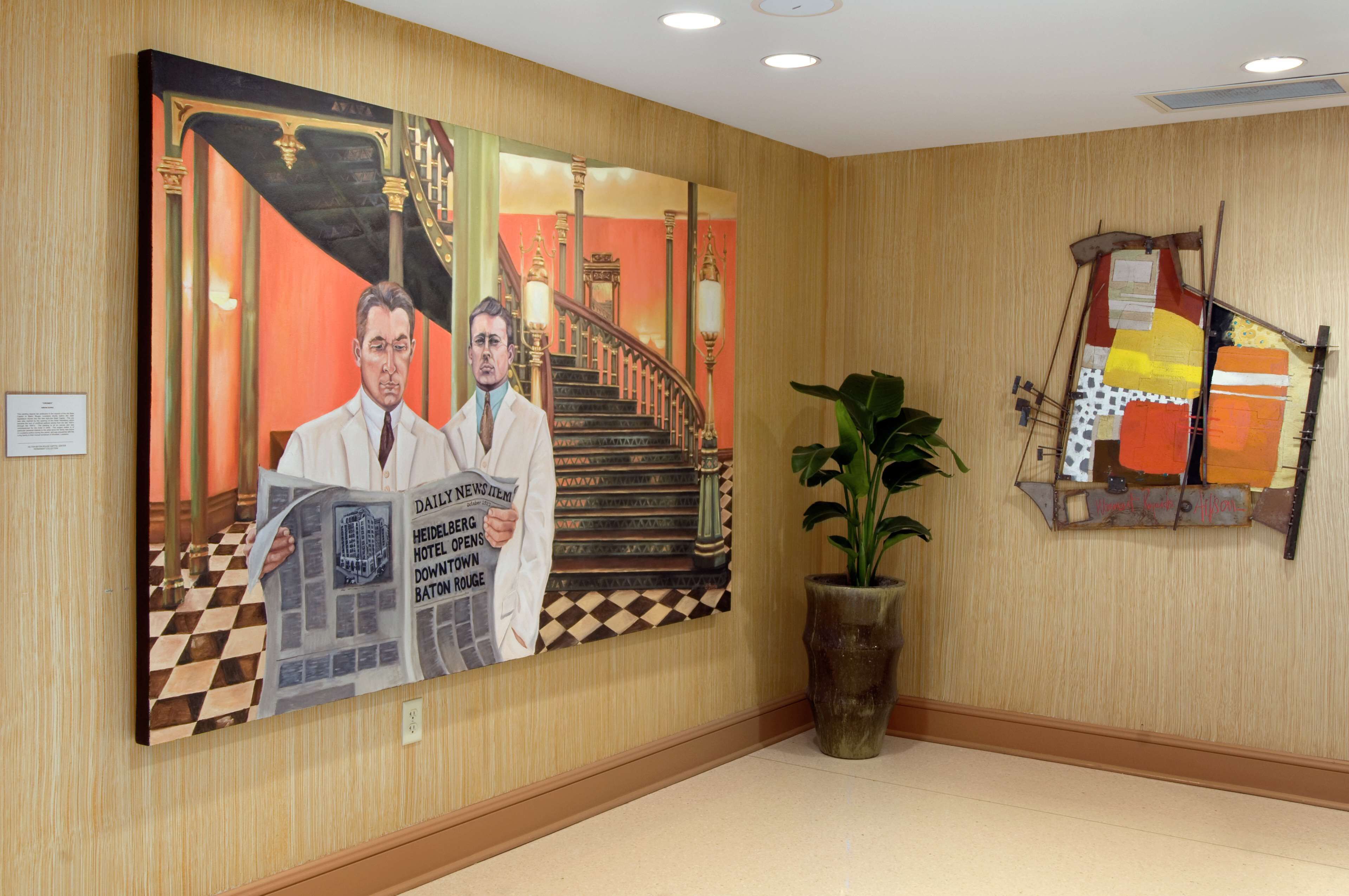 Un hall d’hôtel avec une grande fresque représentant deux hommes en train de lire un journal, un escalier et une plante en pot.