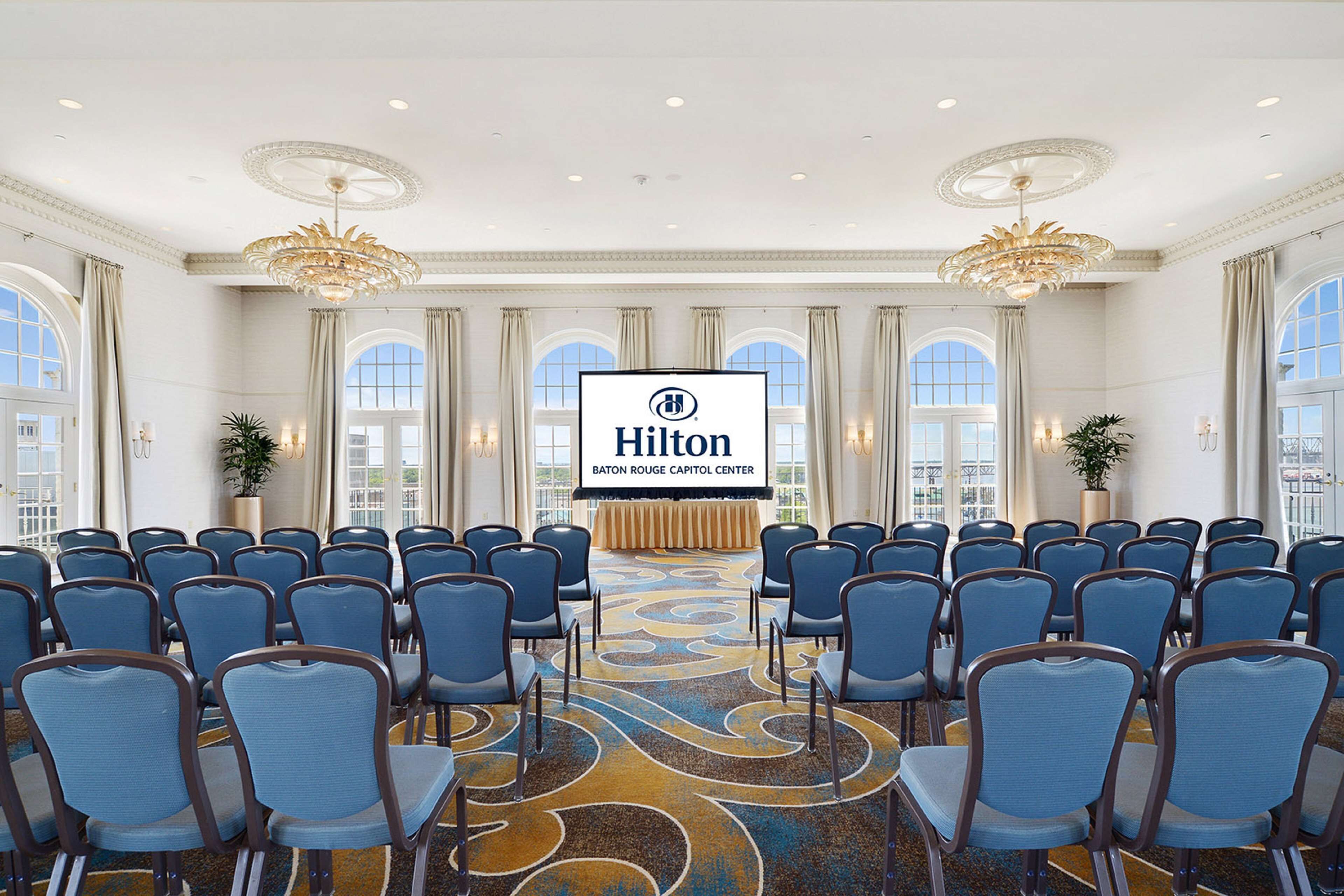 Ein Konferenzraum mit blauen Stuhlreihen, einer großen Leinwand mit dem Hilton-Logo und einem dekorativen Teppich.