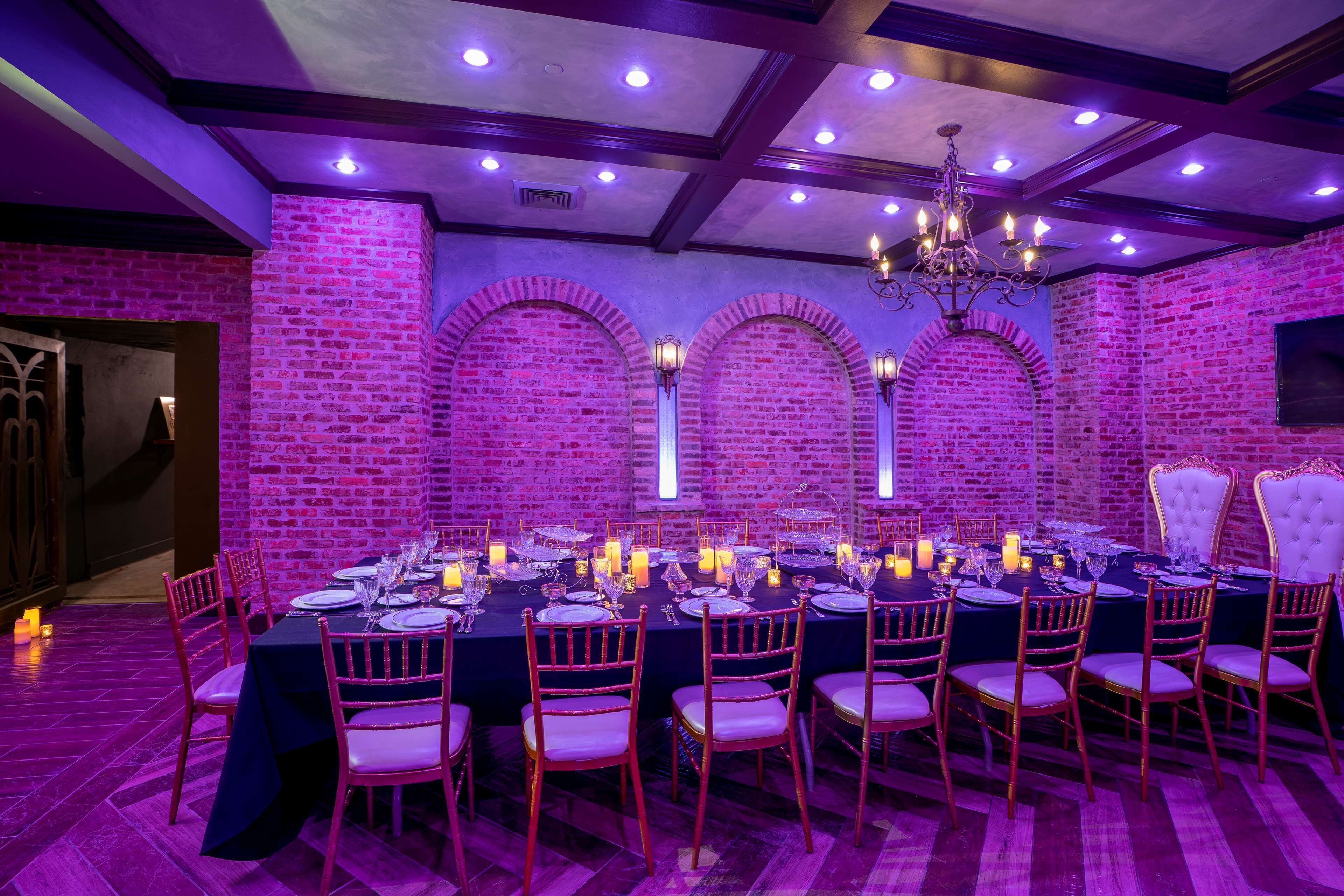 A dining area with a brick wall, arched windows, and a chandelier. Tables are set with candles and silverware.