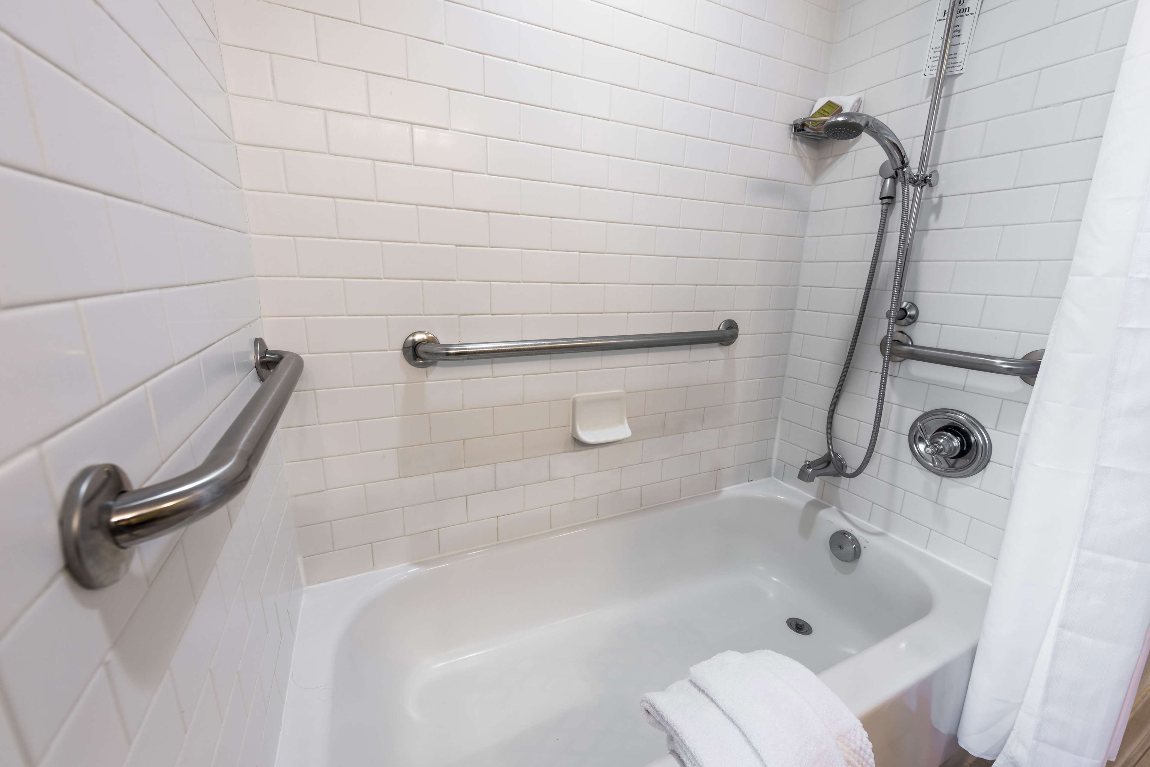 Ein Badezimmer mit weißen Fliesen, einer Badewanne, einer Dusche inklusive Duschbrause und Haltegriffen.