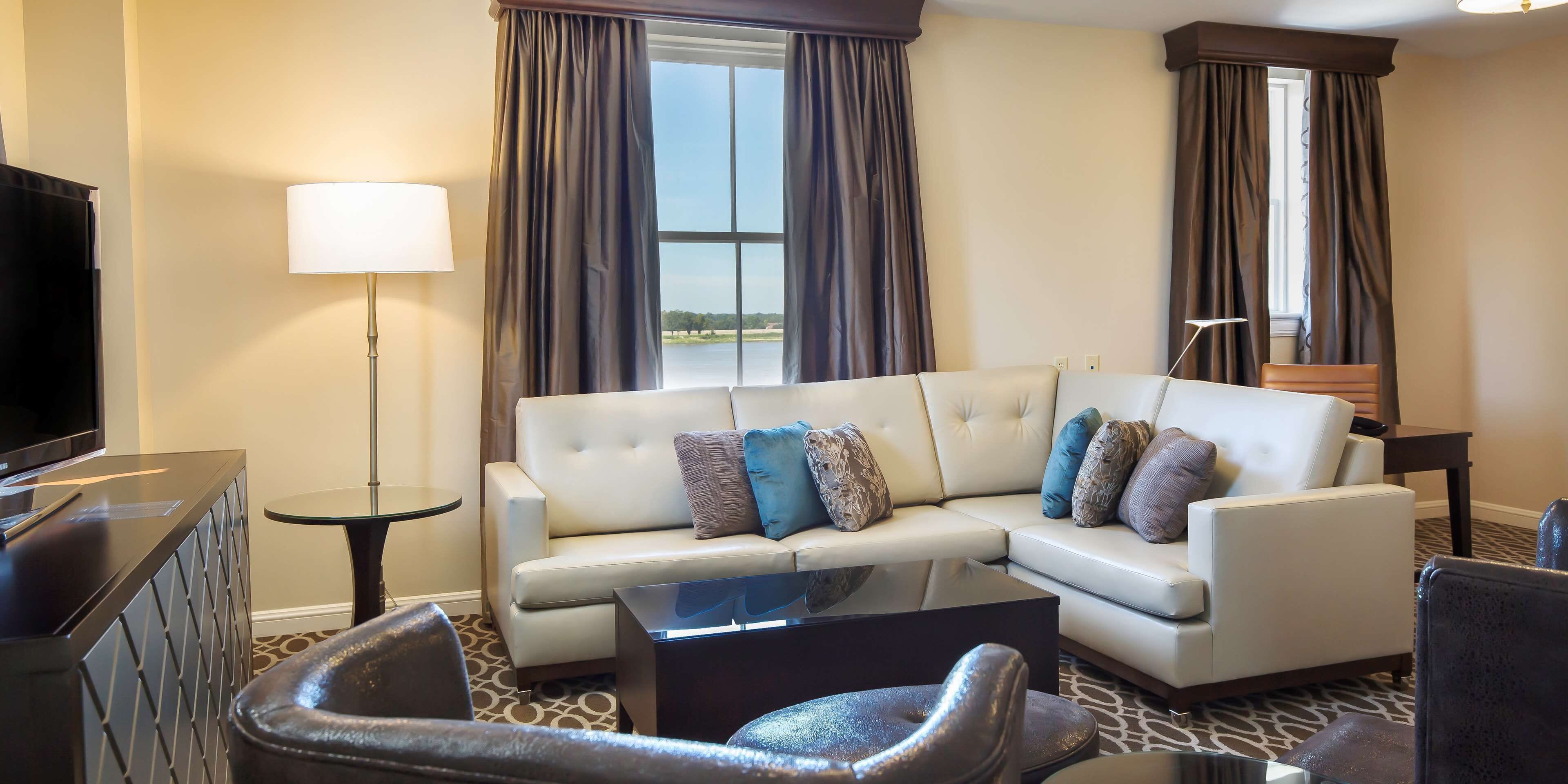 A modern hotel room with a sectional sofa, a flat-screen TV, a glass coffee table, and a view of a body of water through large windows.