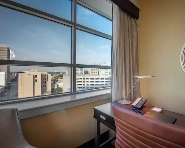 A hotel room with a large window offering a city view, a desk with a lamp, and a telephone.
