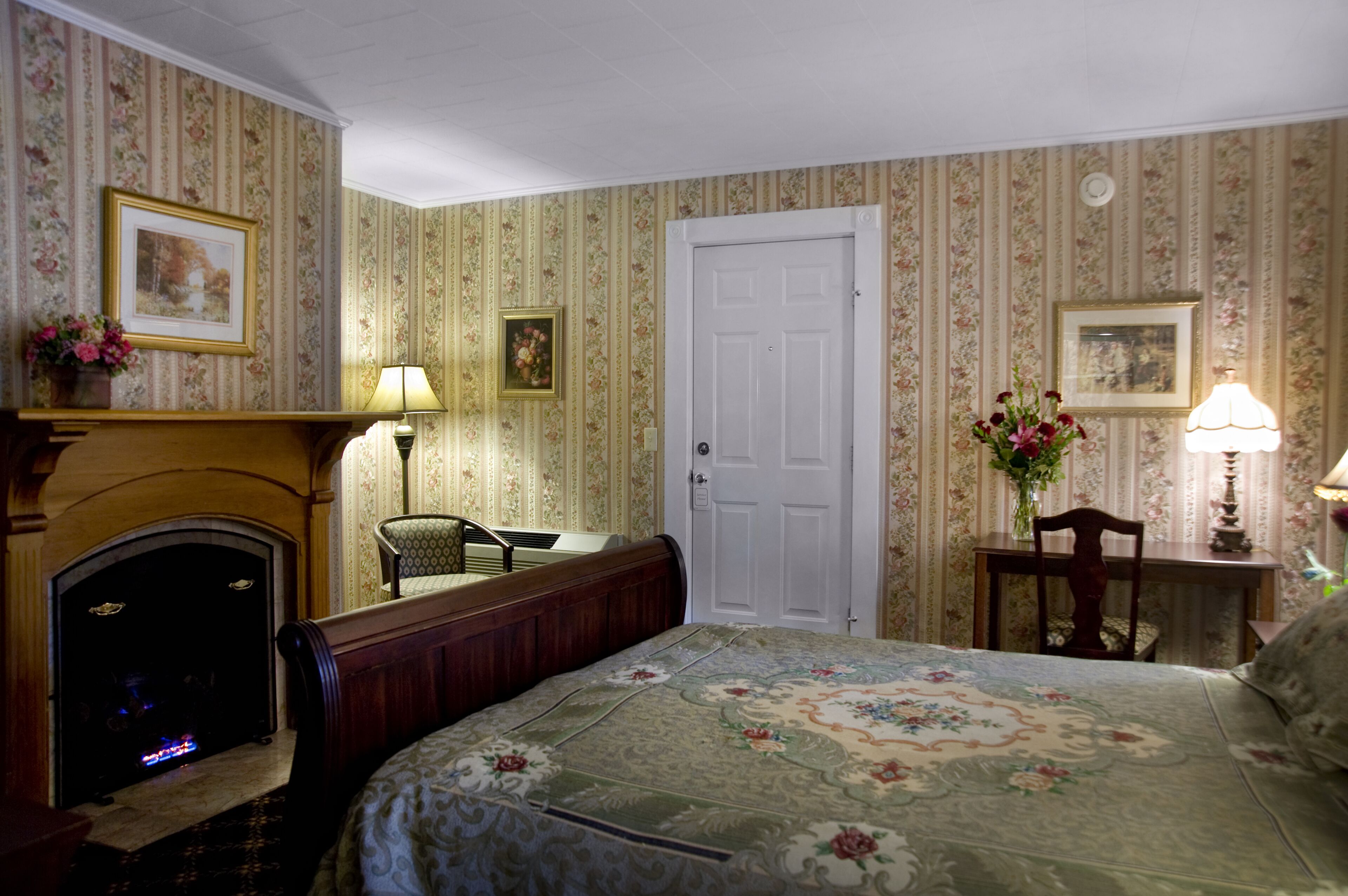 A room with a fireplace, a bed with floral bedding, a chair, a desk with a lamp, and a door.