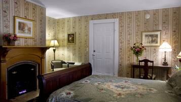 A room with a fireplace, a bed with floral bedding, a chair, a desk with a lamp, and a door.
