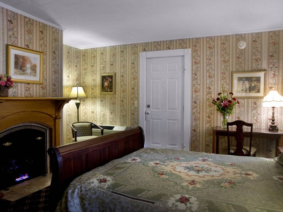 A room with a fireplace, a bed with floral bedding, a chair, a desk with a lamp, and a door.