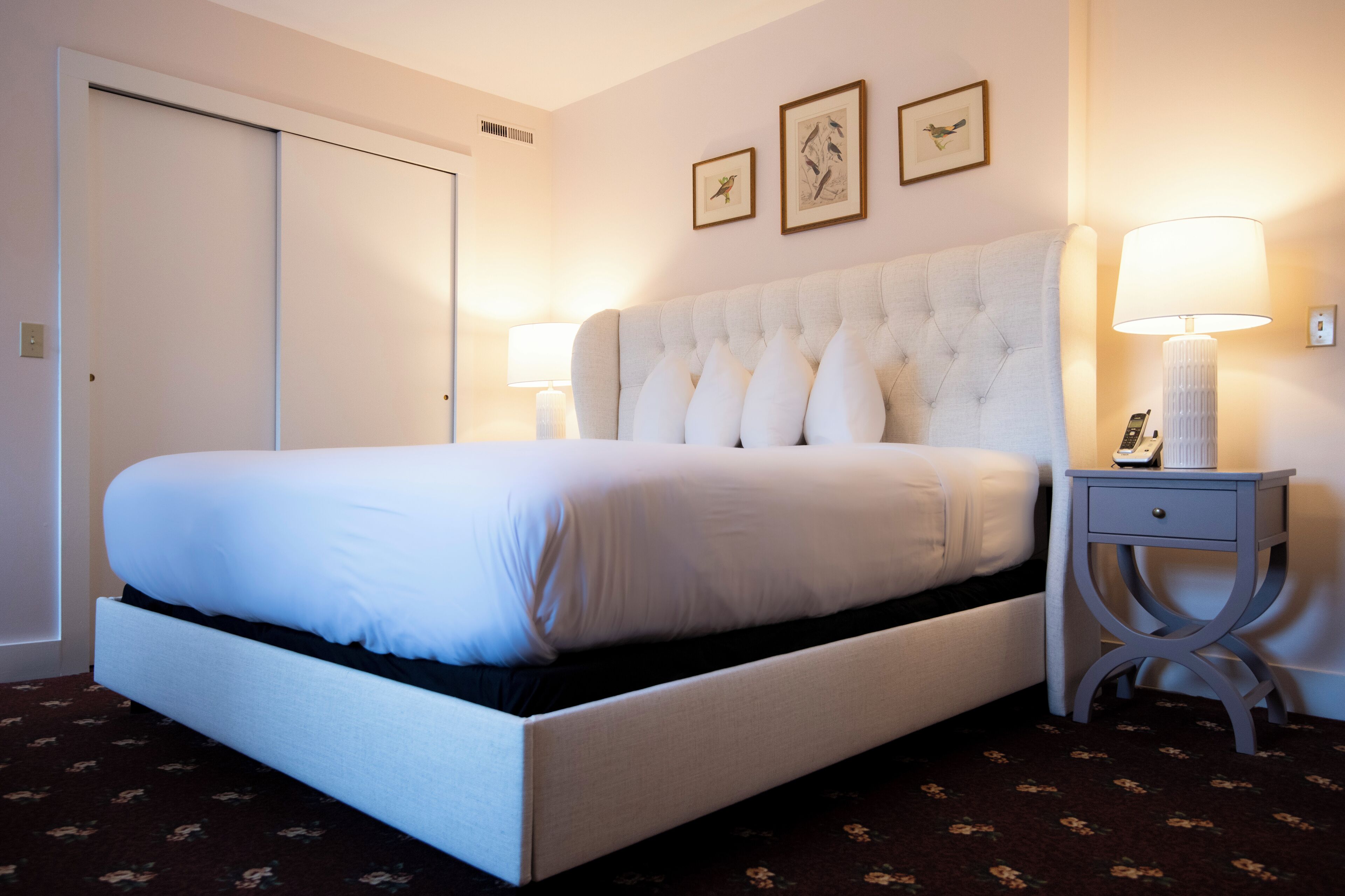 A neatly made bed with a tufted headboard, a bedside table with a lamp, and two framed pictures on the wall.