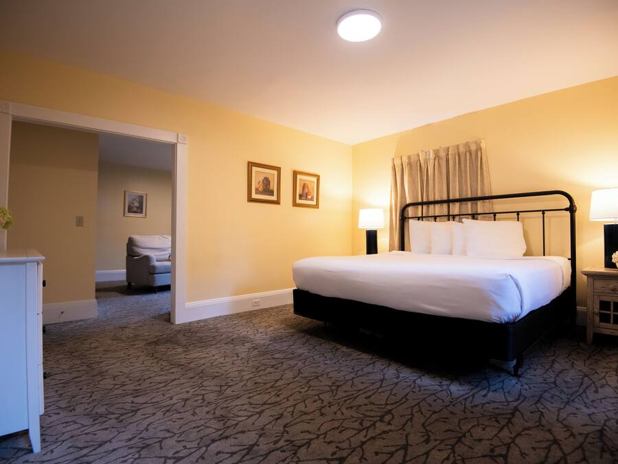 Une chambre d’hôtel avec un grand lit, deux lampes de chevet, une table de chevet avec une lampe, une commode et une porte donnant sur une autre pièce.