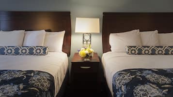 A hotel room with two beds, each with patterned bedding, a nightstand with a lamp and flowers, and a wall-mounted clock.