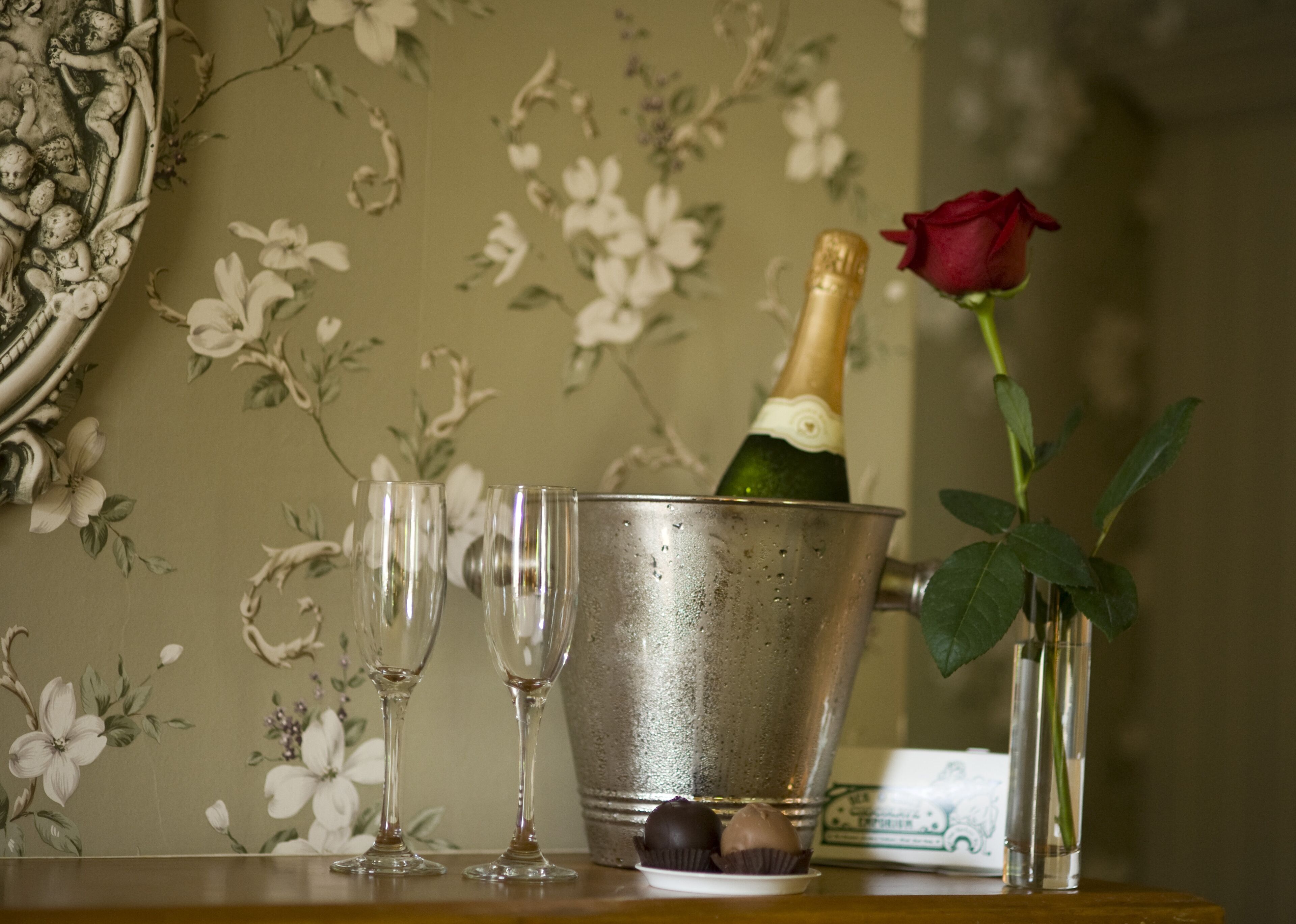 En champagneflaska i en metallhink, två tomma vinglas och en röd ros i en vas på ett bord med en blommig tapet som bakgrund.
