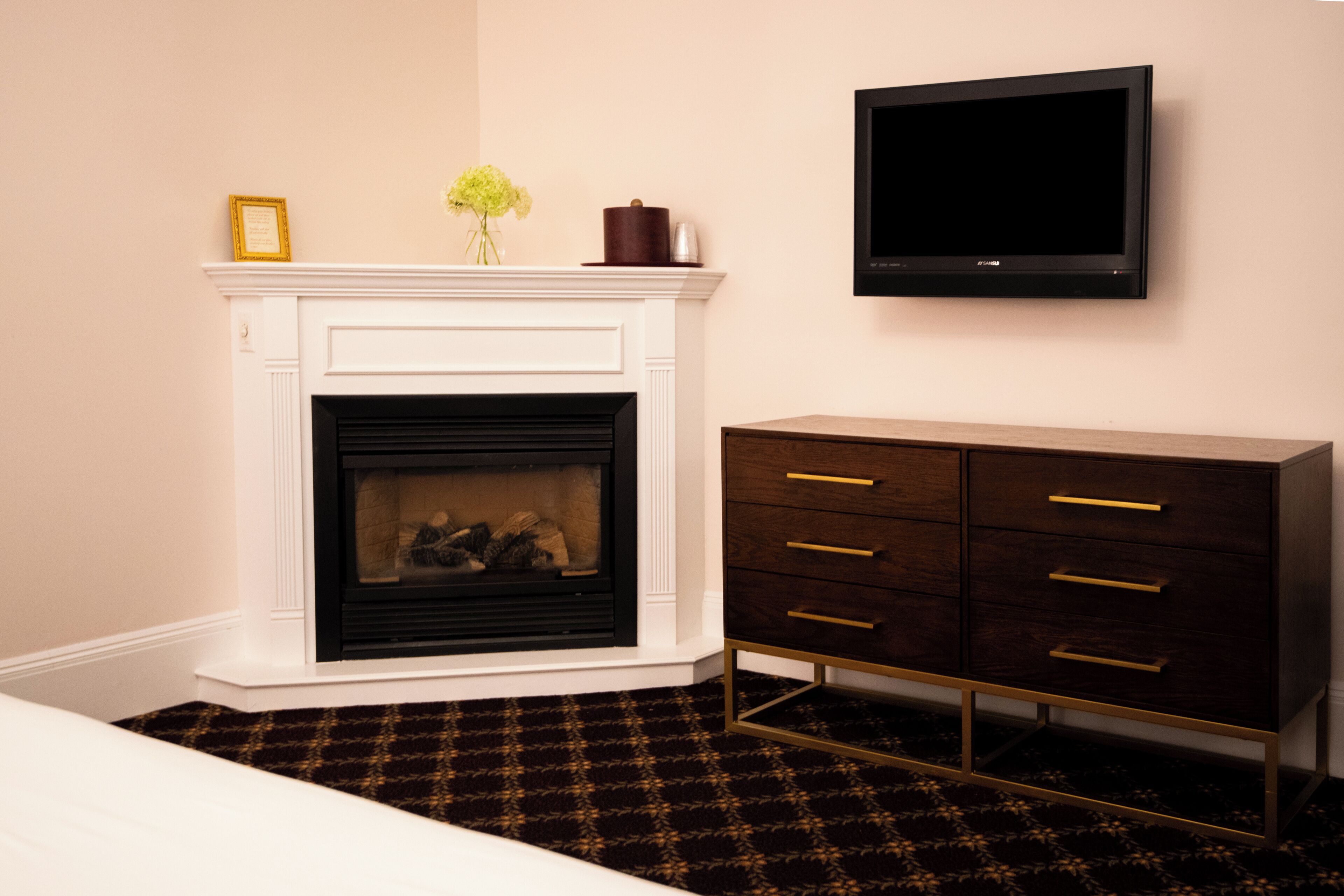 A room with a white fireplace, a dark wooden dresser with gold handles, and a flat-screen TV mounted on the wall.