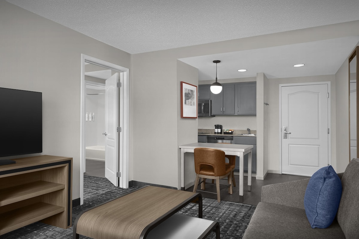A hotel room with a TV, a bed, a sofa, a chair, a desk, a kitchenette, and a bathroom.