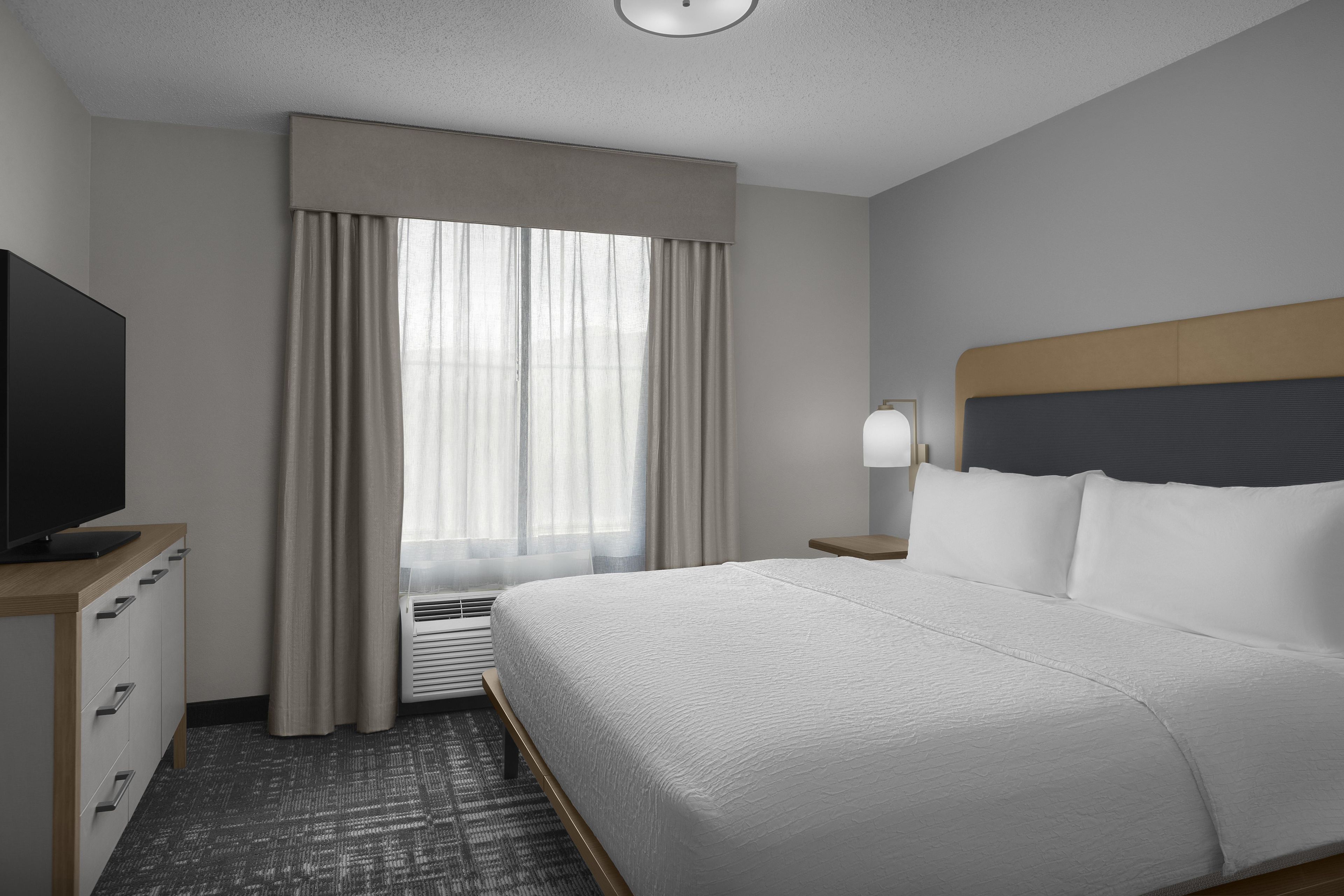 A hotel room with a bed, a television on a wooden cabinet, a window with curtains, and a bedside lamp.