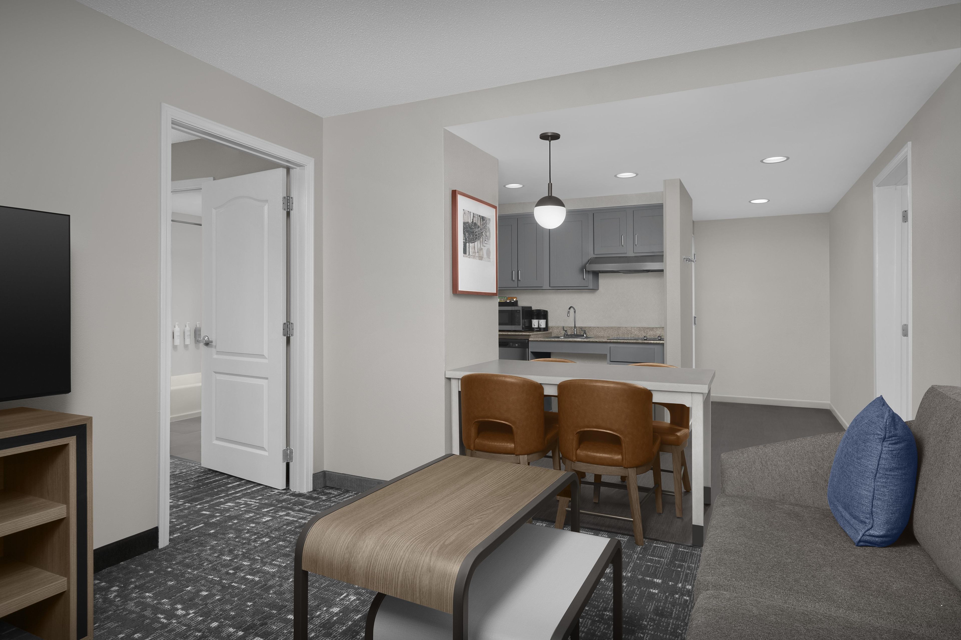 A modern hotel room with a kitchenette, a dining area with a table and chairs, a sofa, and a flat-screen TV.