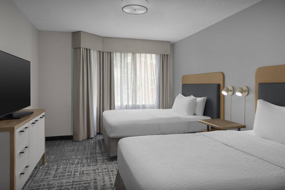 A hotel room with two beds, a TV, a desk, and a window with curtains.