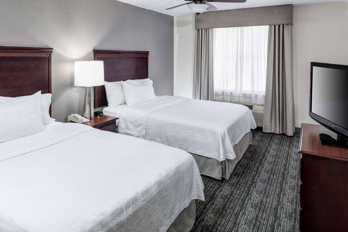 A hotel room with two beds, a desk with a lamp, a TV, and a window with curtains.