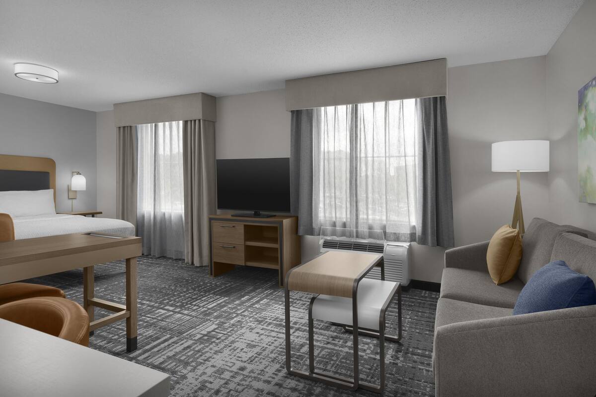 A hotel room with a bed, desk, TV, sofa, and large windows.