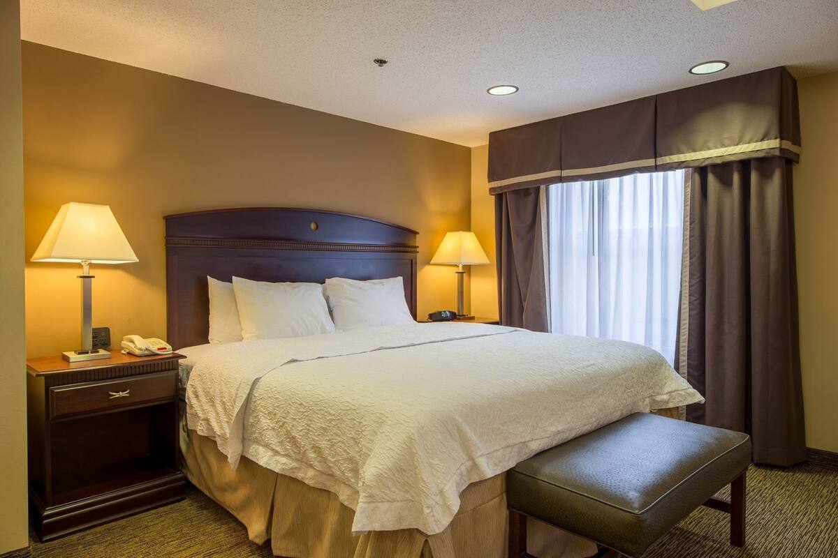 A hotel room with a bed, bedside lamps, a telephone, a bench, and a window with curtains.