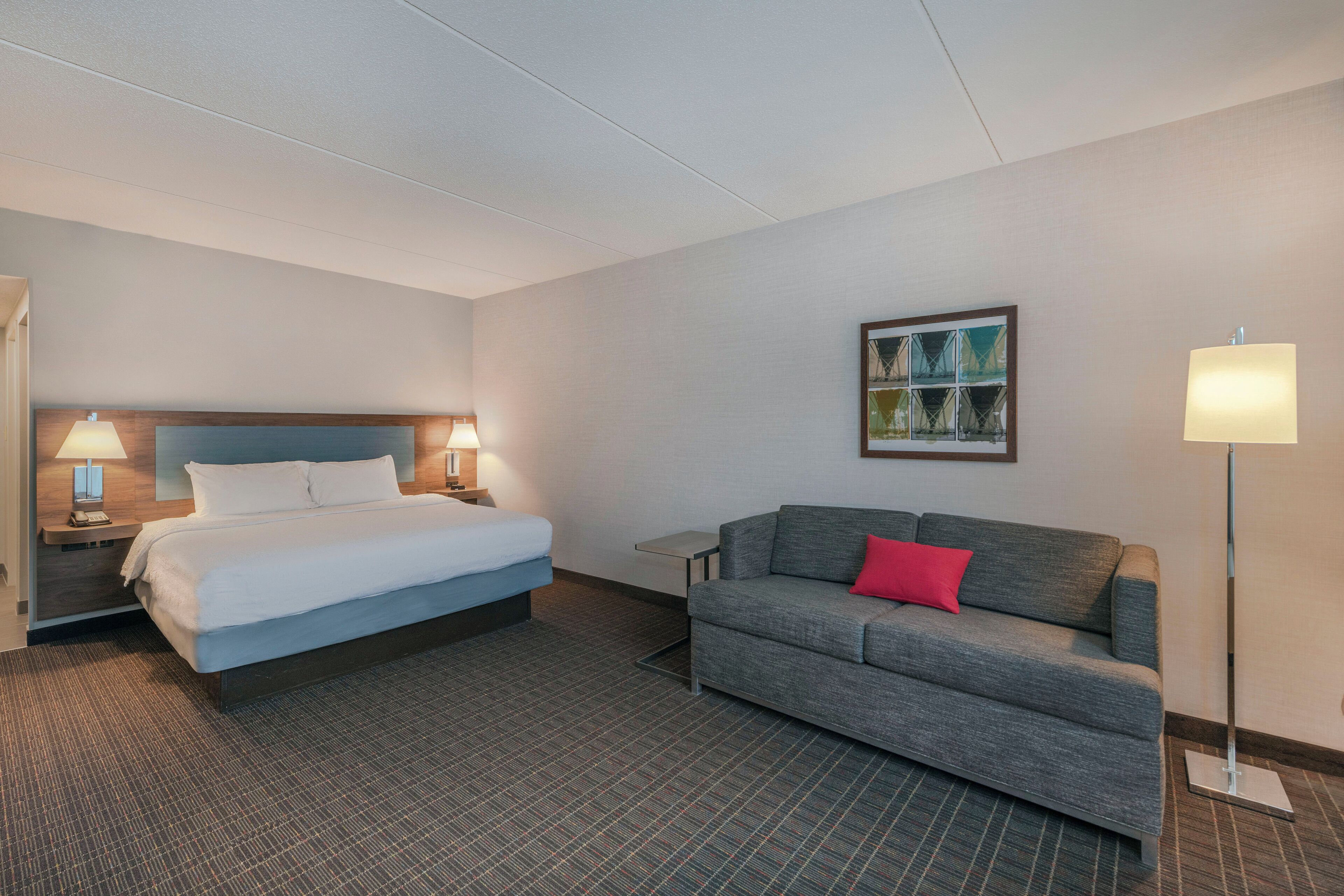 A hotel room with a bed, a grey sofa with a red pillow, a painting on the wall, and a floor lamp.