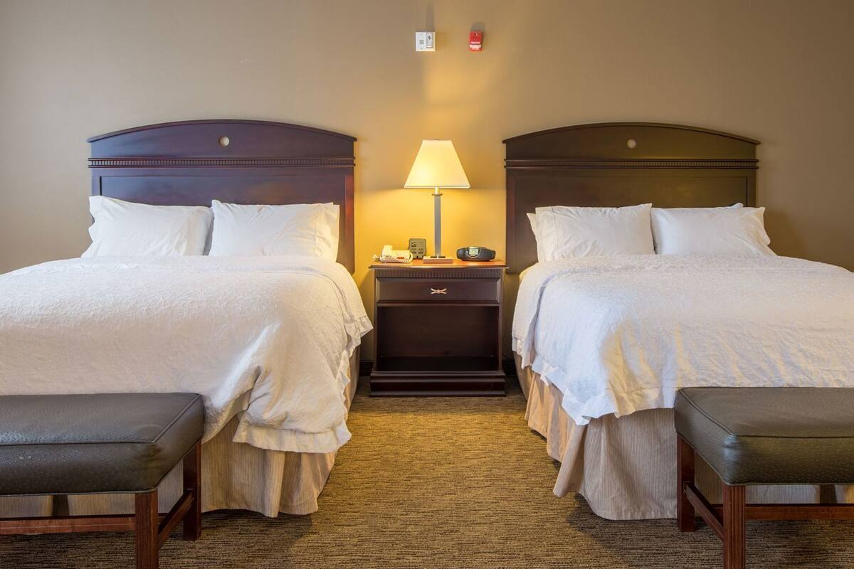 A hotel room with two beds, a nightstand, a lamp, and a bench.