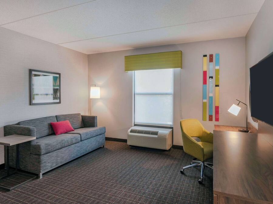 Habitació d'hotel moderna amb un sofà gris, una taula petita, una cadira groga, una televisió de pantalla plana i una finestra gran amb vistes.