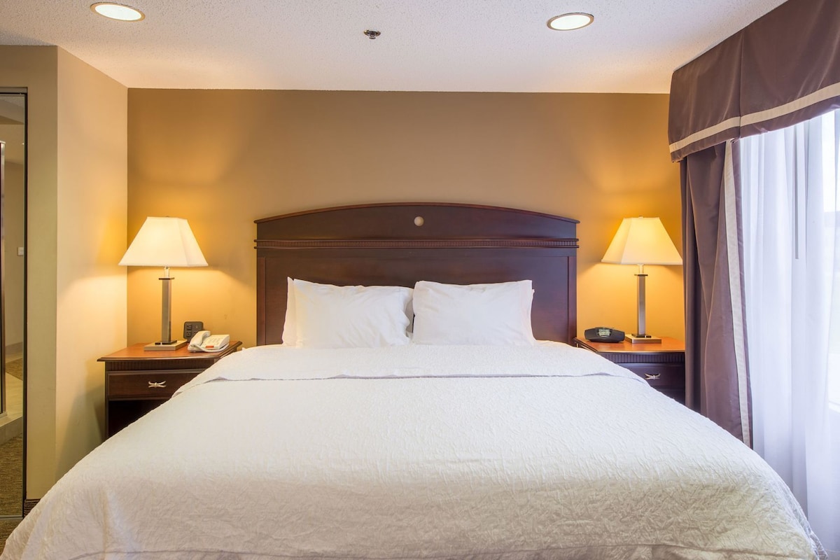 A hotel room with a large bed, two bedside tables with lamps, a telephone, and a window with curtains.