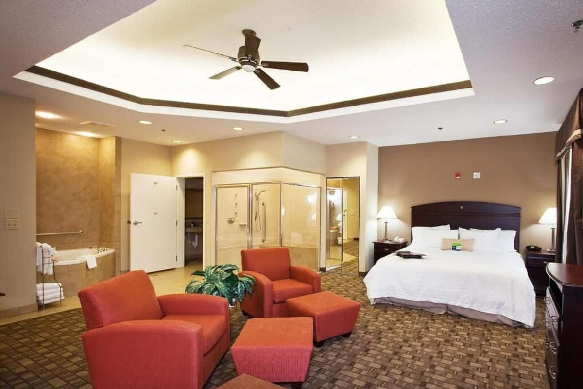 A hotel room with a bed, a ceiling fan, a seating area with chairs and ottomans, a bathroom with a sink and mirror, and a plant.
