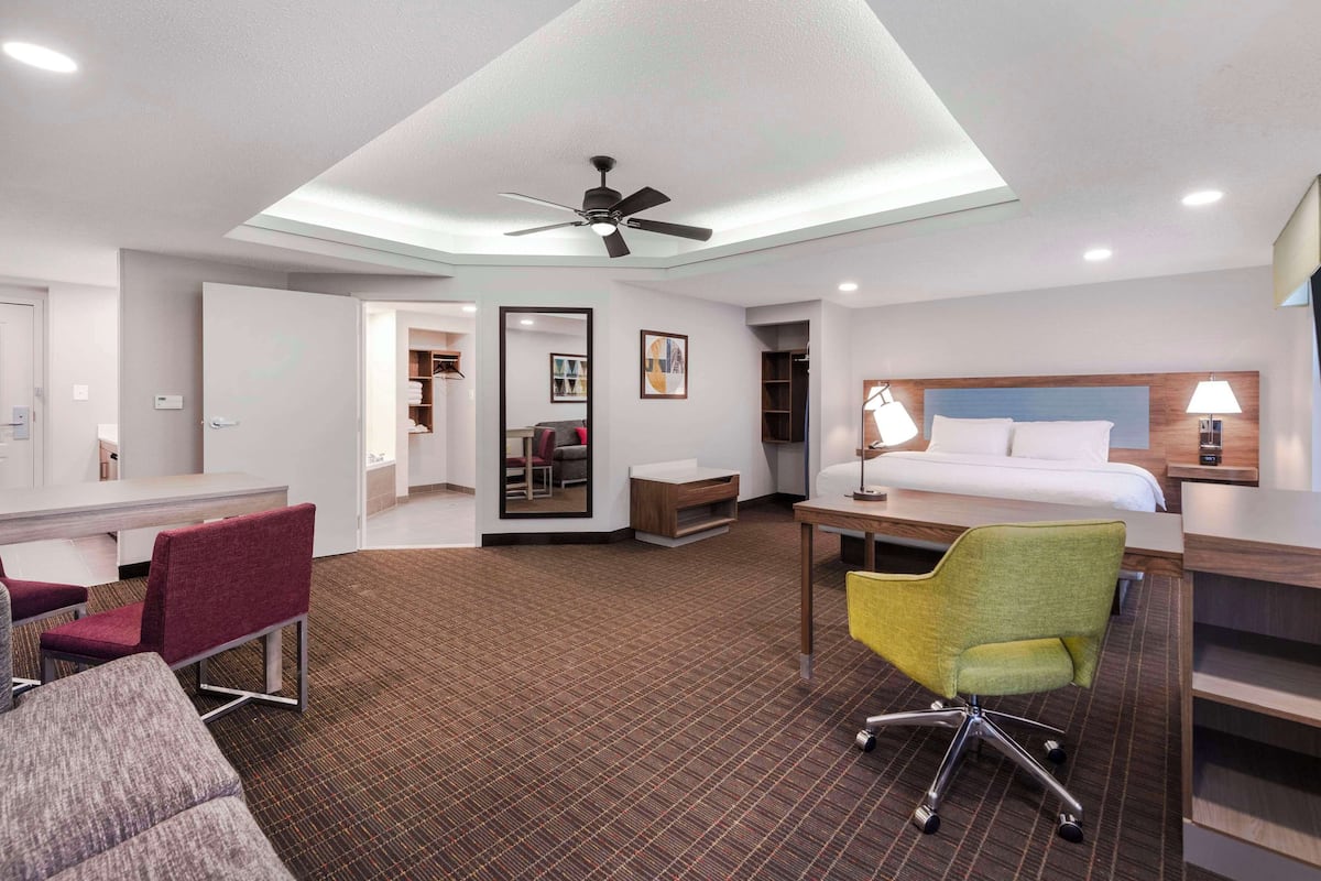 A hotel room with a bed, desk, chairs, a ceiling fan, and a TV.