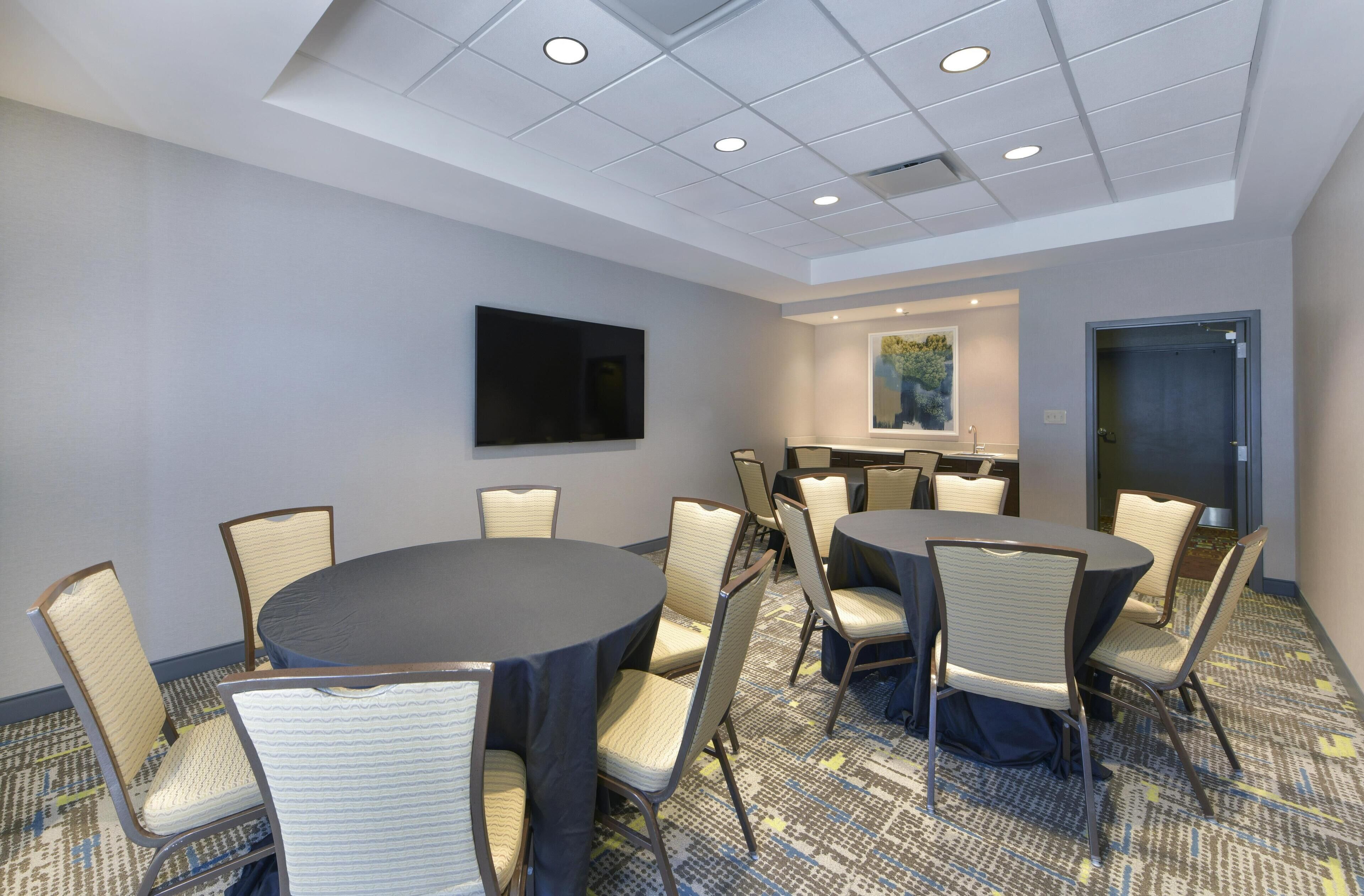 A conference room with round tables and chairs, a flat-screen TV, a painting on the wall, and a door.