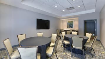 A conference room with round tables and chairs, a flat-screen TV, a painting on the wall, and a door.