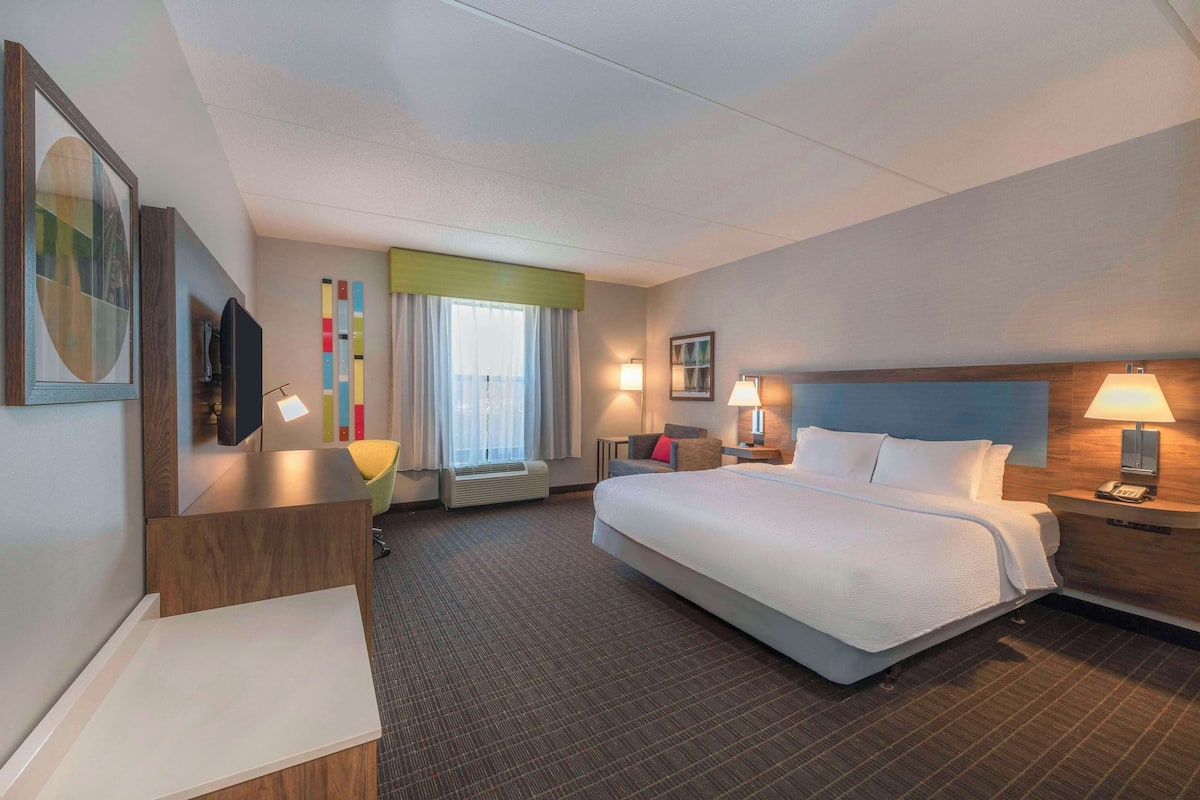 A hotel room with a large bed, a desk, a chair, a TV, a window with curtains, and a wall with a picture and a lamp.