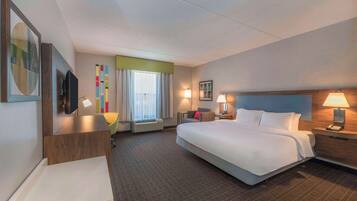 Quarto de hotel com uma cama grande, uma mesa de trabalho, uma cadeira, uma televisão, uma janela com cortinas e um quadro com uma luminária na parede.