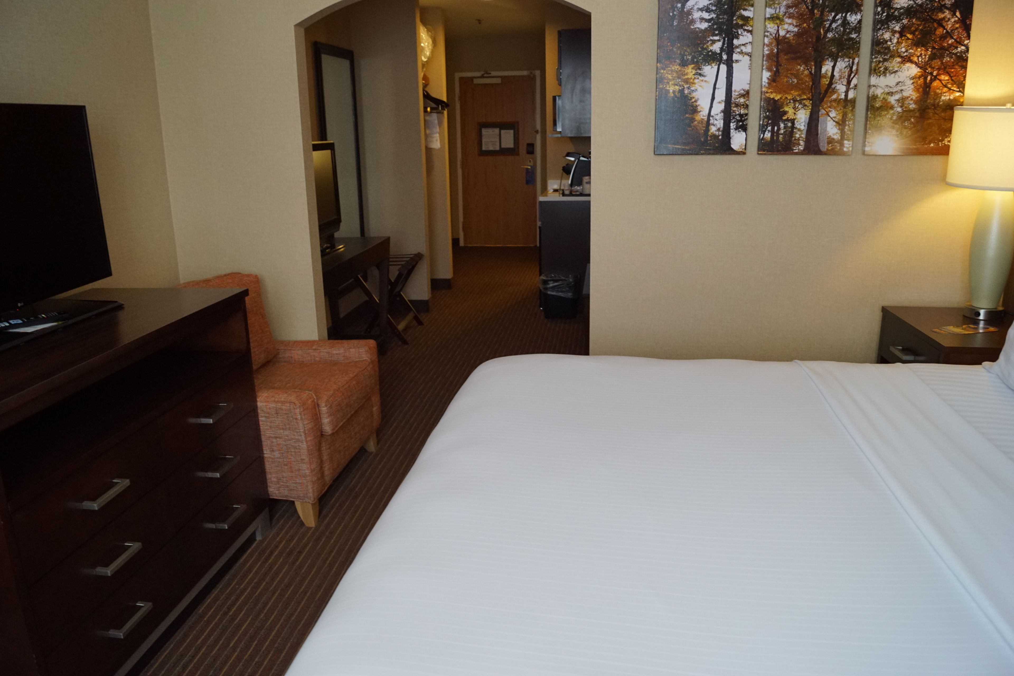 A hotel room with a bed, a desk with a television, a chair, a desk lamp, and a painting on the wall.