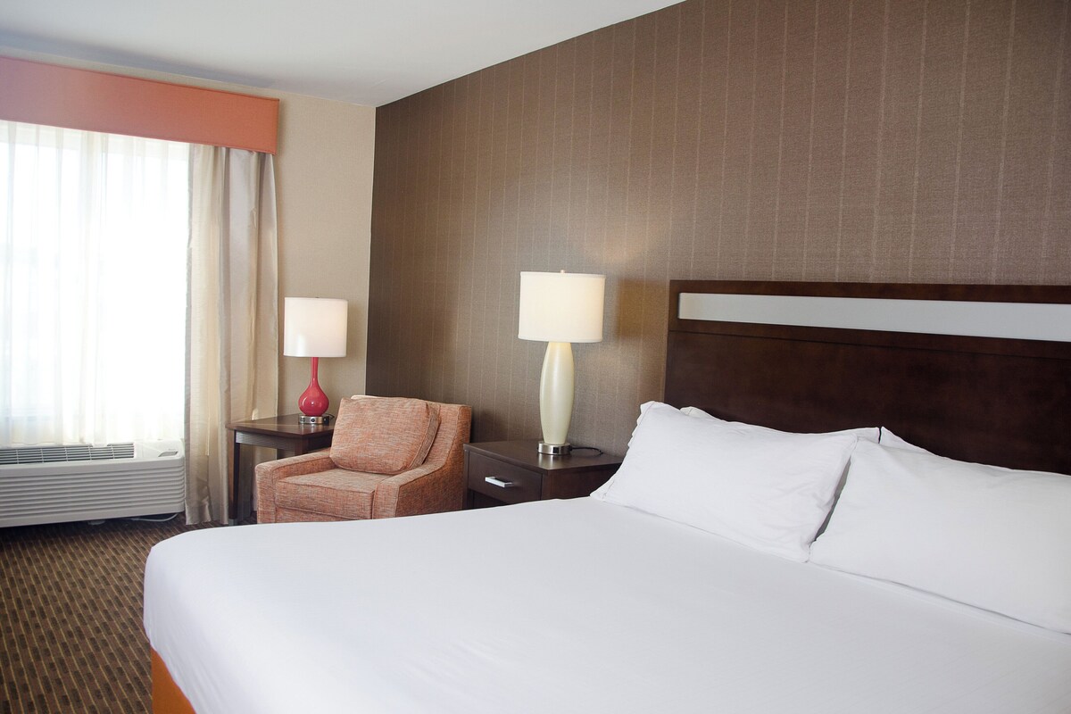 Habitación de hotel con una cama grande, una silla, dos mesitas de noche con lámparas y un ventanal con cortinas.