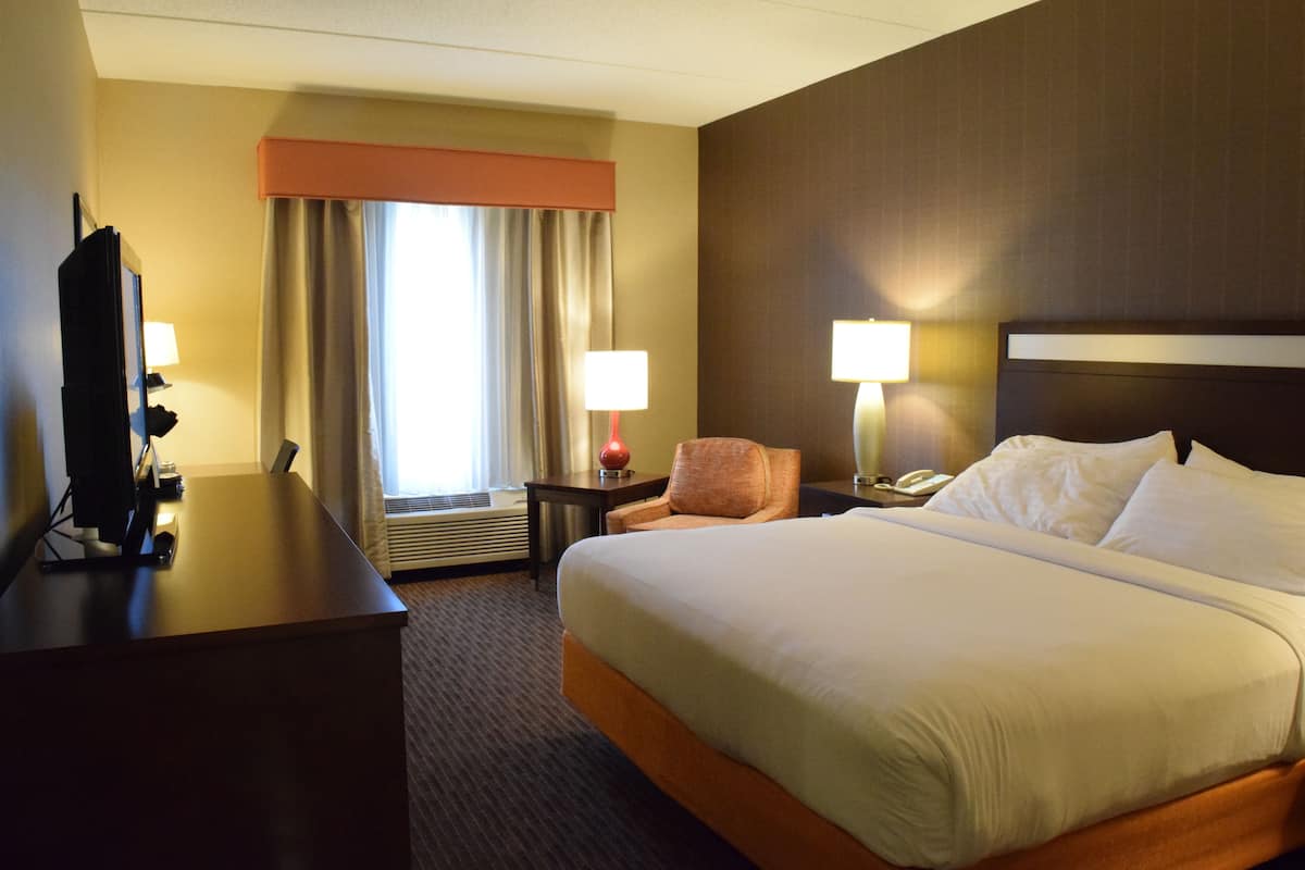 Habitación de hotel con una cama grande, un escritorio con computadora, una silla, dos lámparas de noche y un ventanal con cortinas.