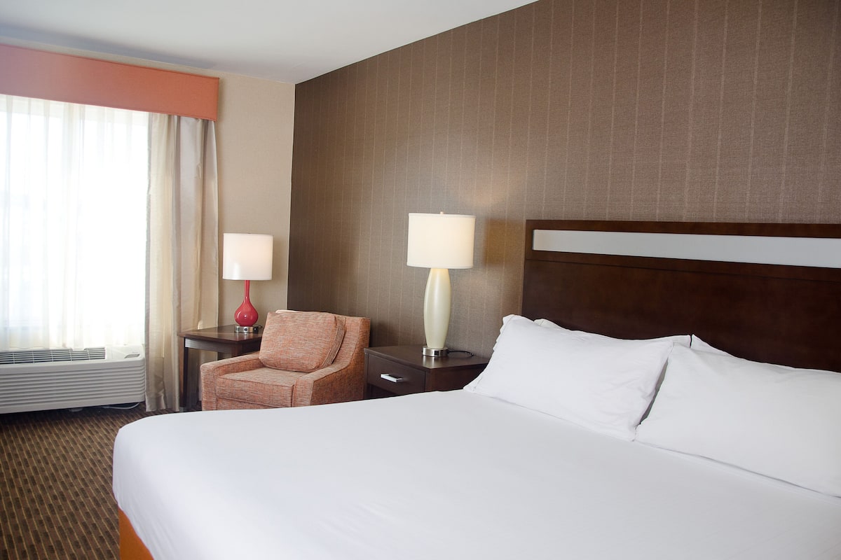 Habitación de hotel con una cama grande, una silla, dos mesitas de noche con lámparas y un ventanal con cortinas.