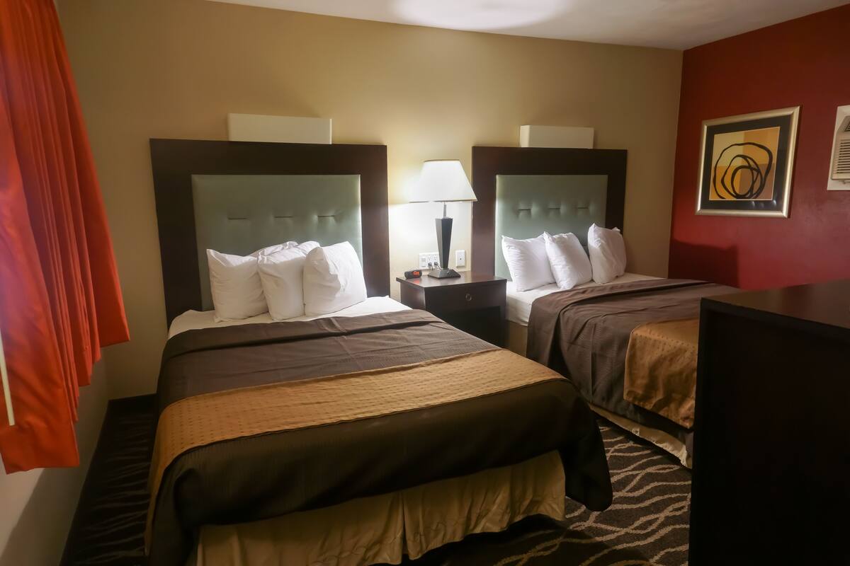 A hotel room with two beds, a nightstand with a lamp, and a red wall with a framed abstract painting.