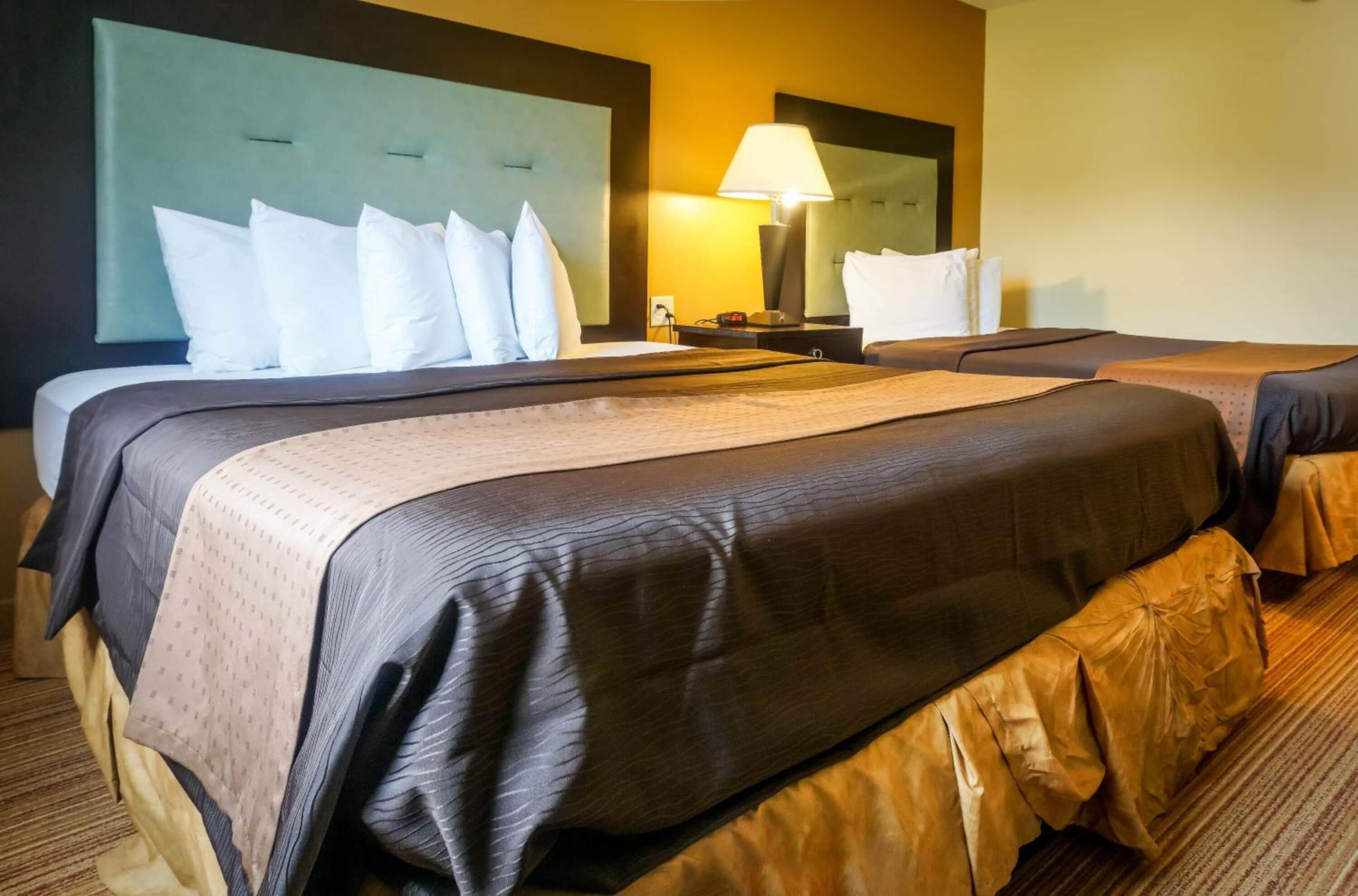 A hotel room with two beds, a headboard, a nightstand with a lamp, and a large mirror.