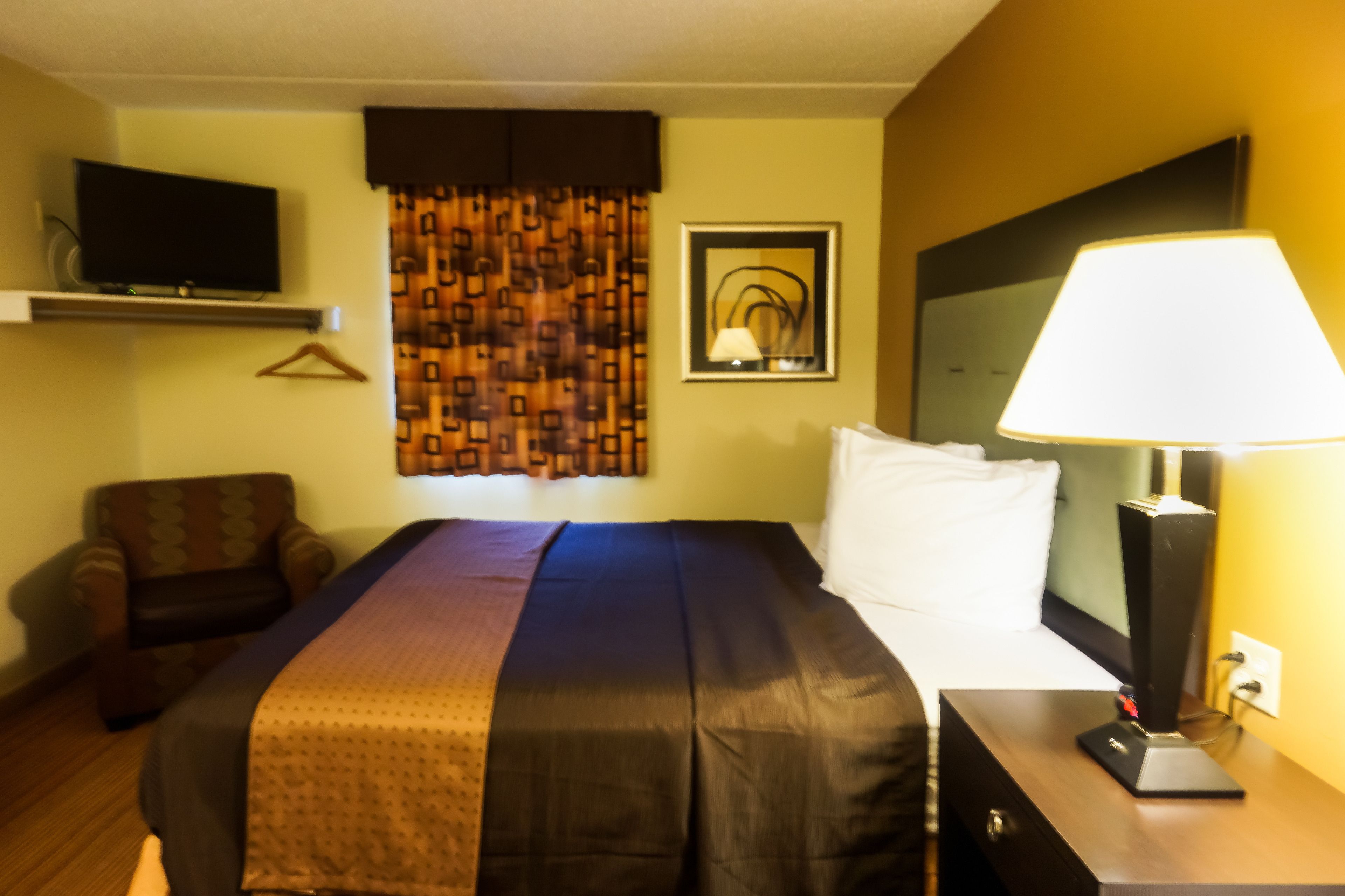 A hotel room with a bed, a TV mounted on the wall, a lamp on a bedside table, and a chair.