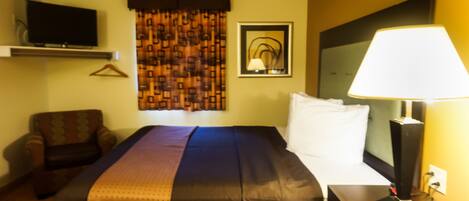 Habitación de hotel con una cama, un televisor montado en la pared, una lámpara en una mesita de noche y una silla.