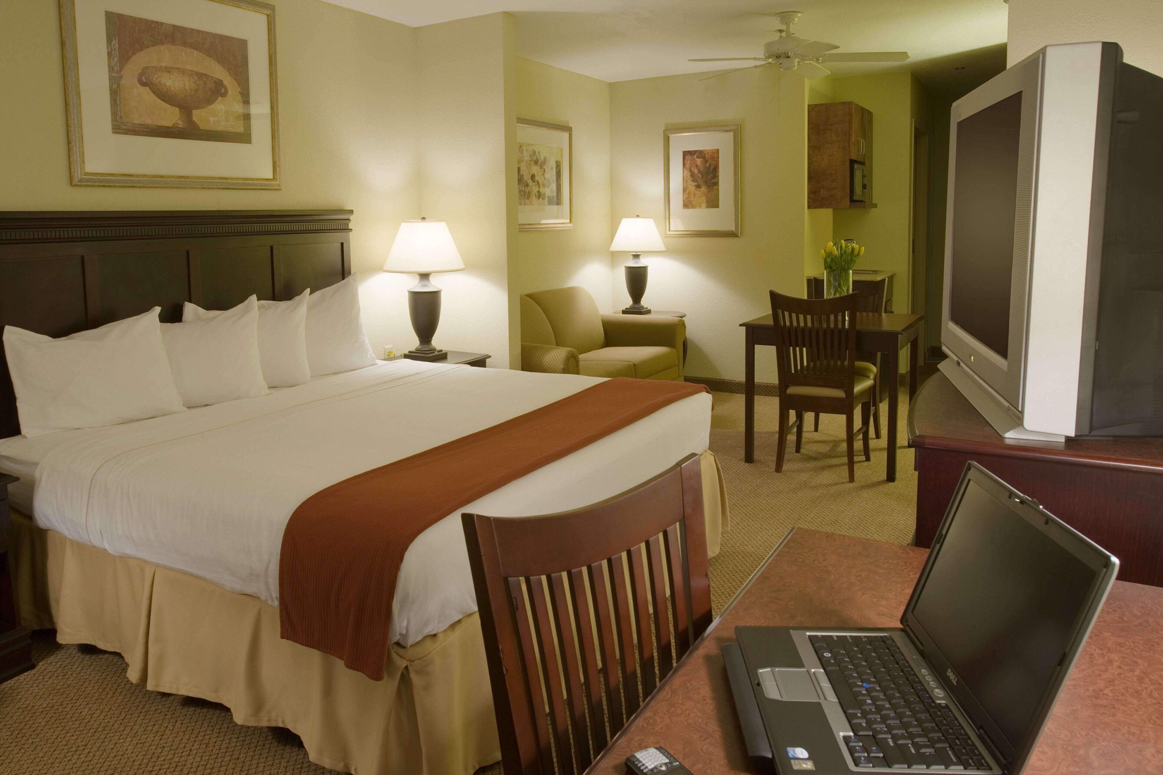 A hotel room with a bed, a desk with a laptop, a television, and a dining area with chairs and a table.
