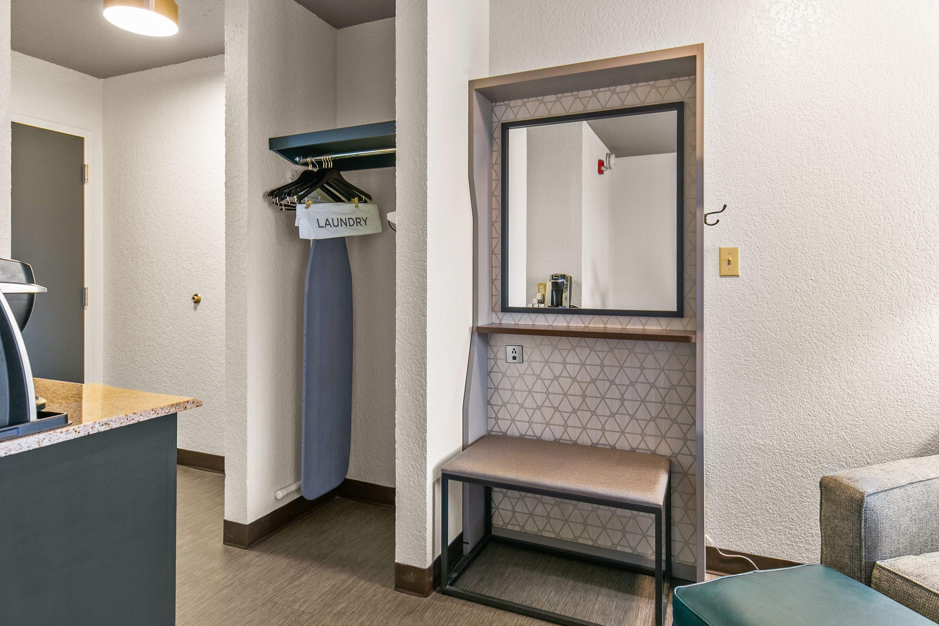 Una habitación de hotel con cama, una mesita, un banco, un espejo, un estante en la pared y un cartel de lavandería.
