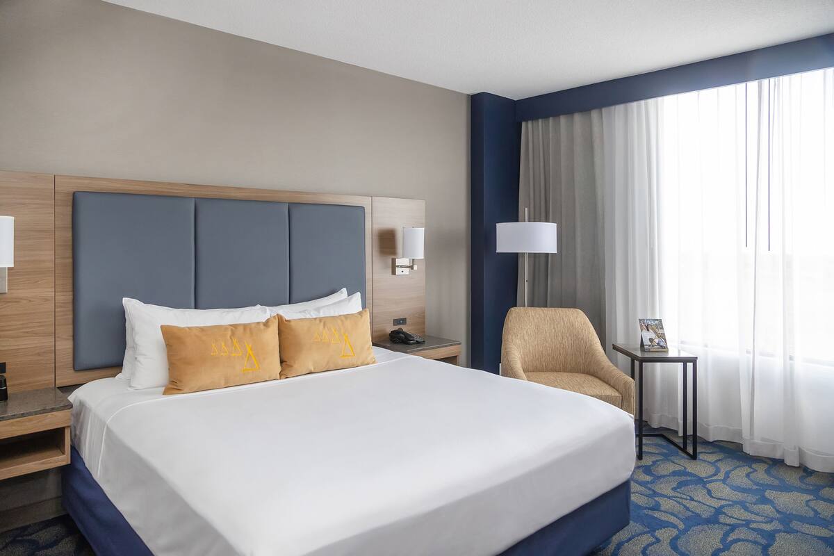 Una habitación de hotel con una cama grande, una silla, una mesita de noche, una lámpara y una ventana con cortinas.
