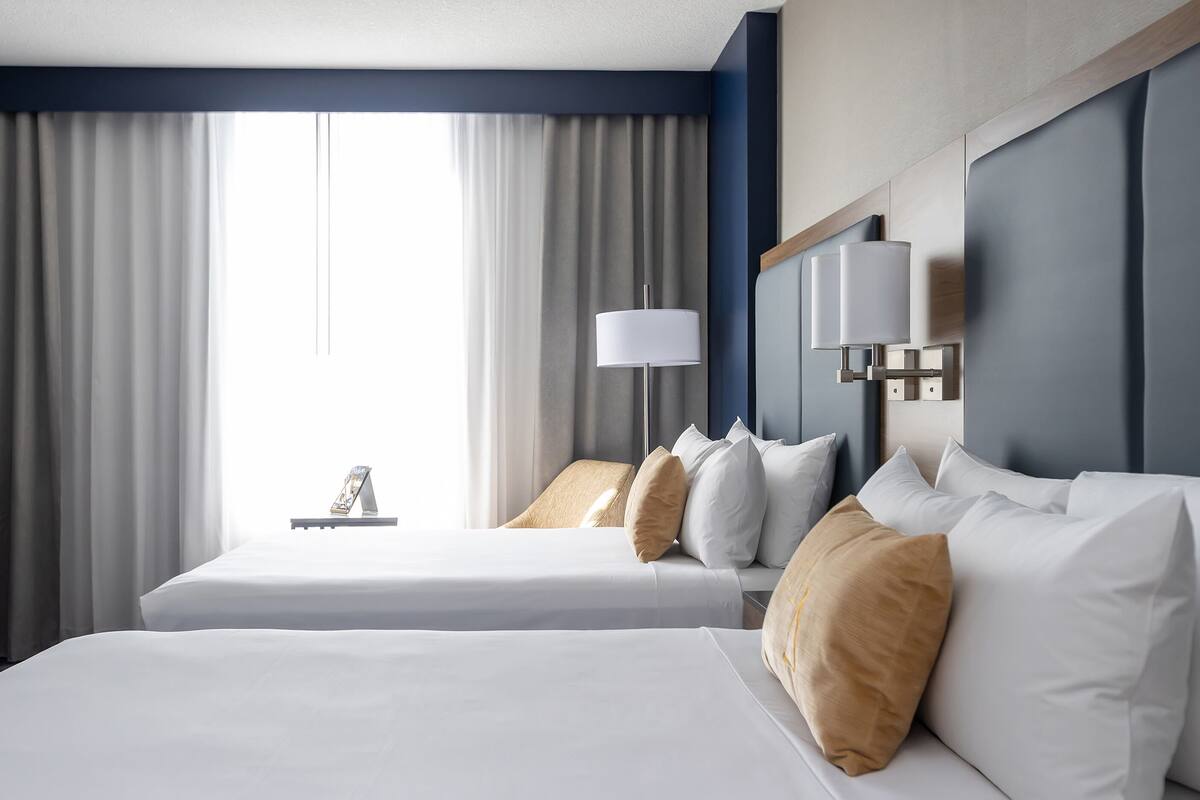 Habitación de hotel con dos camas, un ventanal grande con cortinas, una mesita de noche con lámpara y un cabecero moderno.