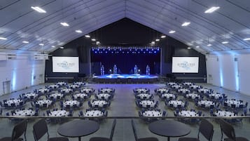 Un grand espace intérieur doté d’une scène et de plusieurs rangées de tables et de chaises, probablement utilisé pour des événements ou des conférences.