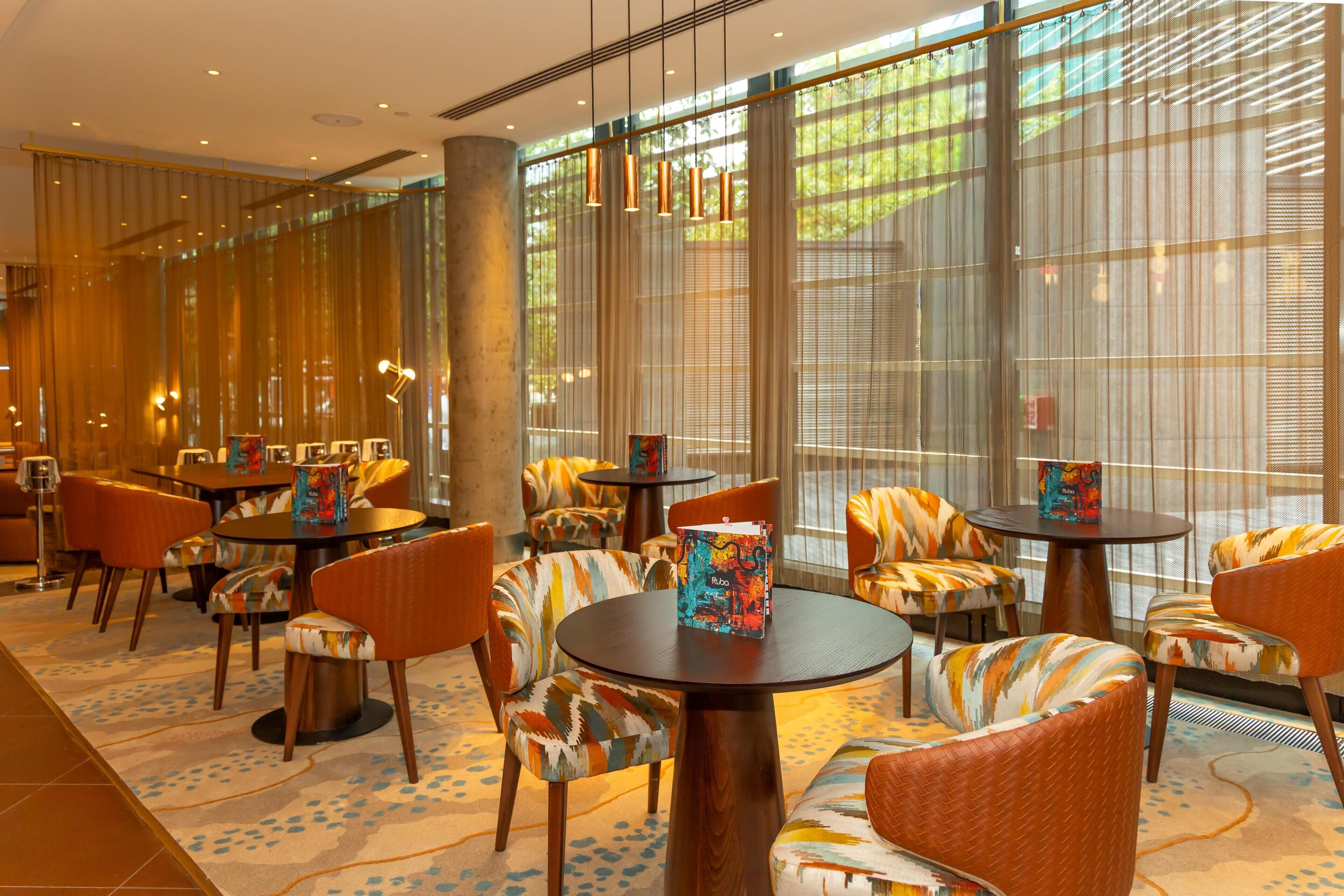 A hotel lobby with round tables, patterned chairs, and large windows with sheer curtains.