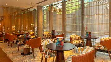 A hotel lobby with round tables, patterned chairs, and large windows with sheer curtains.